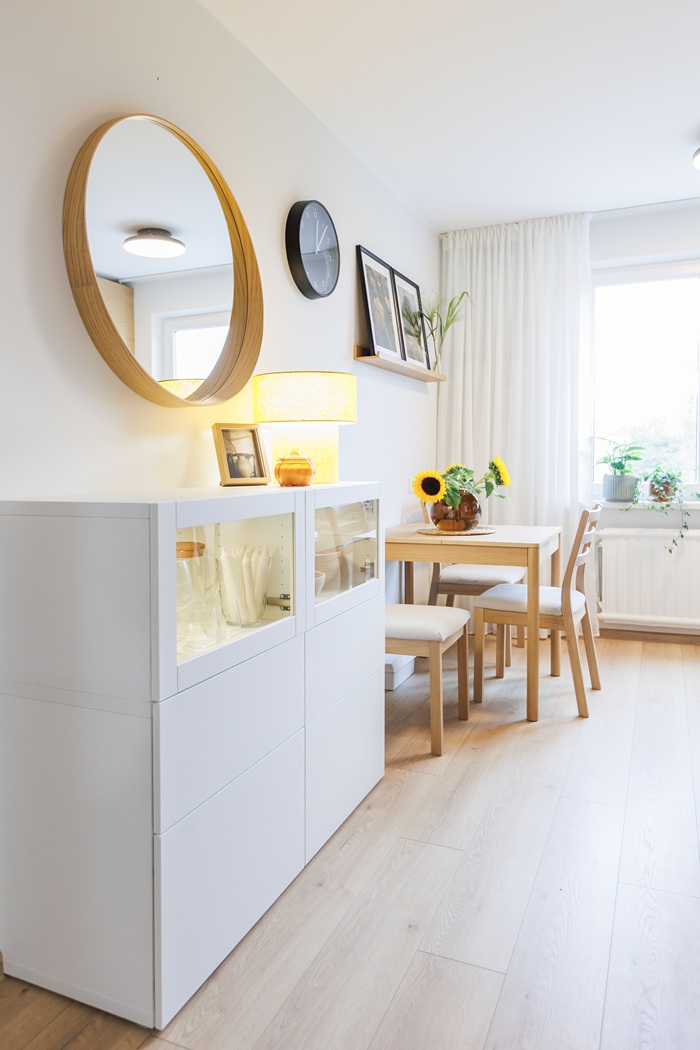 Šviesus valgomasis su nedideliu mediniu stalu, dviem kėdėmis, balta indauja ir apvaliu veidrodžiu ant sienos.