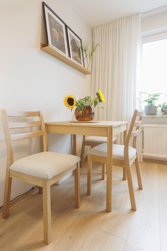 Vaizdas iš arti: medinis valgomojo stalas su dviem kėdėmis, vazoje pamerktomis saulėgrąžomis ir įrėmintomis nuotraukomis ant sieninės lentynos.