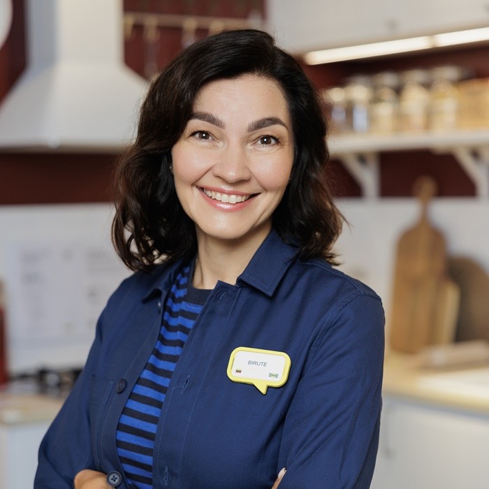 IKEA interjero dizainerė su mėlynos spalvos uniforma stovi modernioje virtuvės ekspozicijoje ir šypsosi.