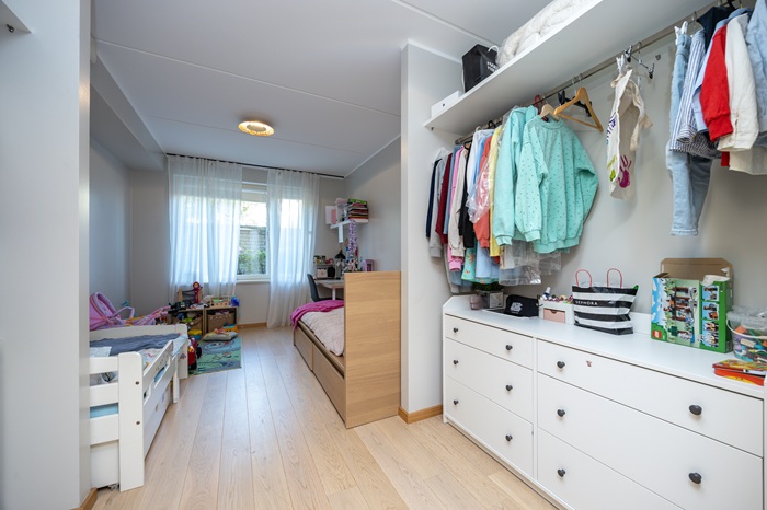 Piklik heledates toonides lastetuba. Esiplaanil paremas servas on valge kummut ja selle kohal ripuvad riidepuutorul värvilised riided. Tagapool on voodid.