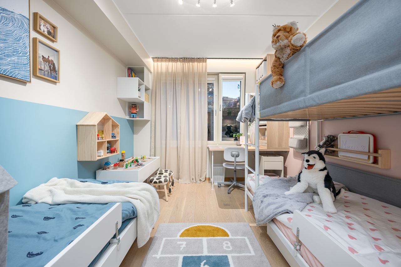 Rahulik lastetuba, kus seinte valget värvi täiendavad mahedad roosad ja sinised toonid. Seinte ääres on voodi ja narivoodi, toa otsaseinas on akna all vasakul mängulaud ja paremal kirjutuslaua ja tooliga õppimisnurk.