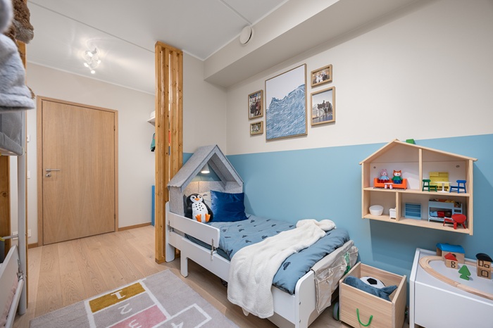 Heledates toonides lastetuba, kus pooleldi valge ja pooleldi beebisinise seina ees on sinise voodipesuga voodi. Voodijalutsis on mänguasjakast ja toa seinal on mööblit täis nukumaja. Garderoobiosa on kaugemas seinas.