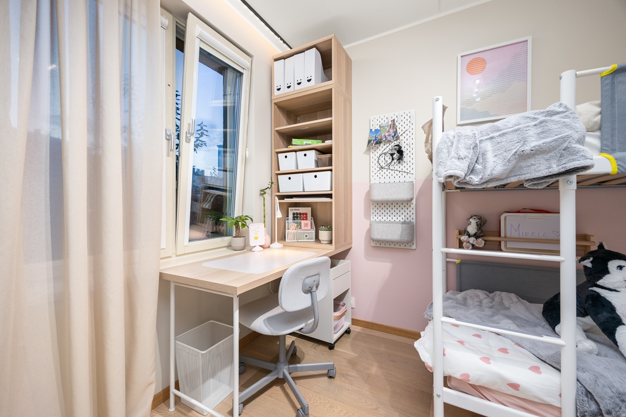 Roosa seinaga õpinurk, mille kõrval paistab valge narivoodi ots. Õpinurgas seisab akna all valge kirjutuslaud, riiul ja valge kirjutuslauatool.