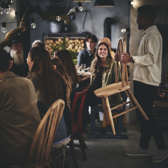 A group of people socialising around the table, with one person holding a wooden chair, about to join them.