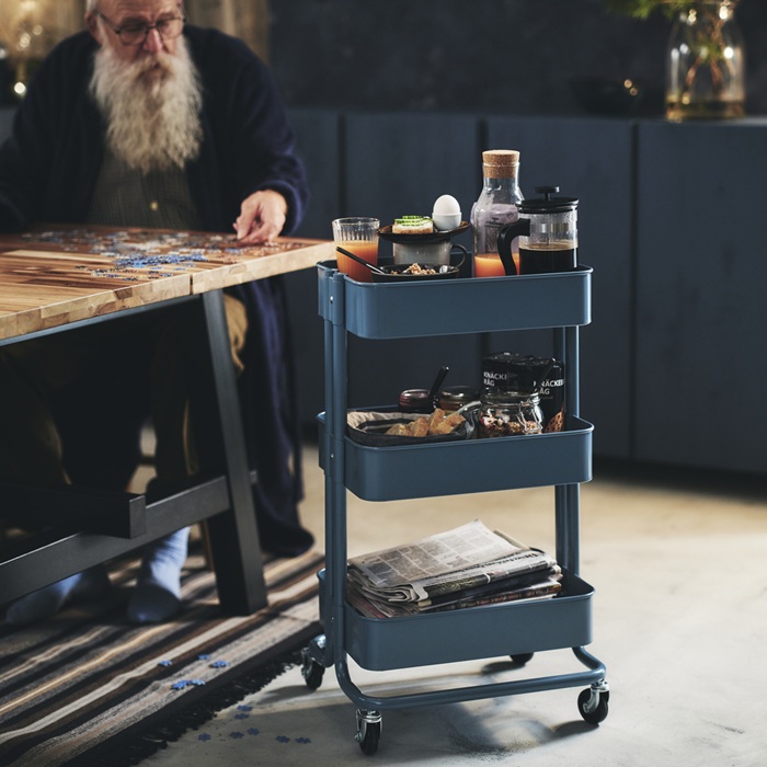 An elderly person assembling a puzzle at a wooden table, with a blue trolley holding drinks, snacks and newspapers nearby.