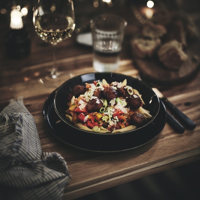 A dark GLADELIG deep plate filled with meatballs and pasta, set on a wooden table.
