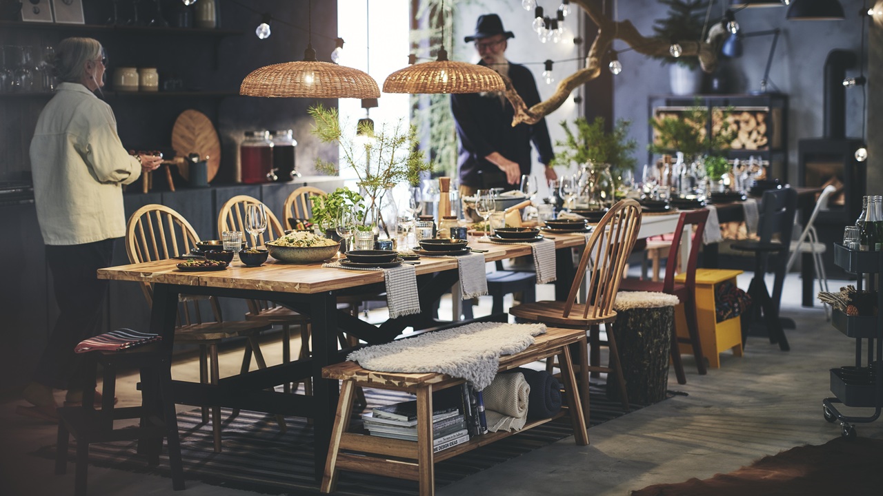 A cosy dining room with a wooden table set for a meal, surrounded by mismatched chairs and warm lighting. Two people are standing nearby.