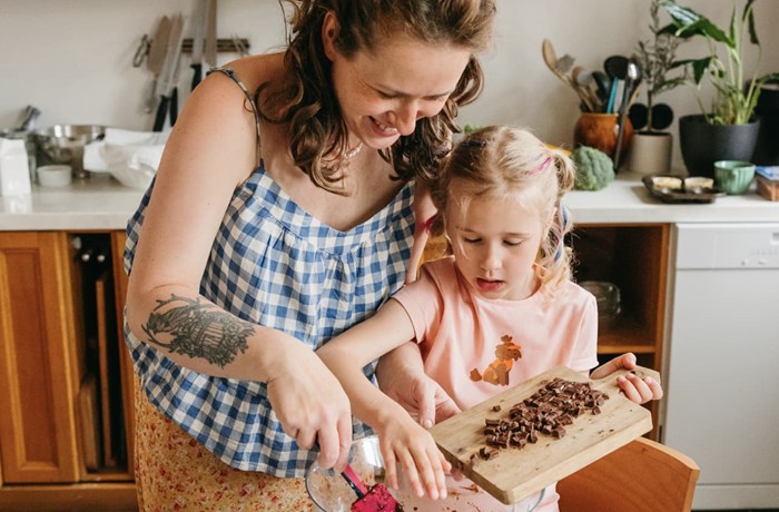 A mother and daughter are working in the kitchen. The girl is pouring chopped chocolate pieces from a wooden board into a glass bowl, while her mother helps her.
