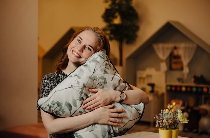 A smiling woman against a background, where a toy kitchen is located, hugging and resting her head on a pillow with a floral print.