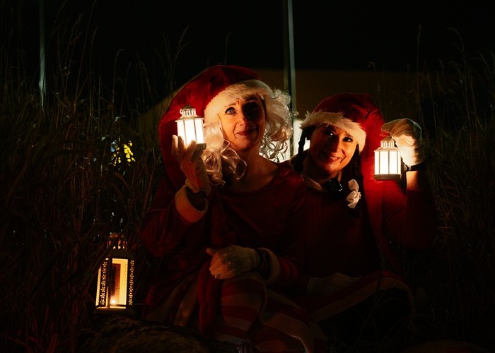 Двое эльфа, одетых в красное, стоят в темноте, держа в руках фонари, которые отбрасывают теплый свет на их лица.