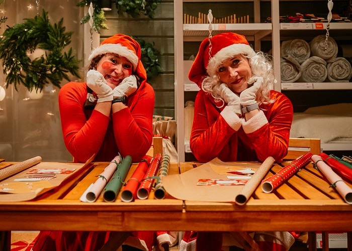 Двое веселых эльфов сидят за рабочим столом, покрытым разноцветными рулонами упаковочной бумаги.