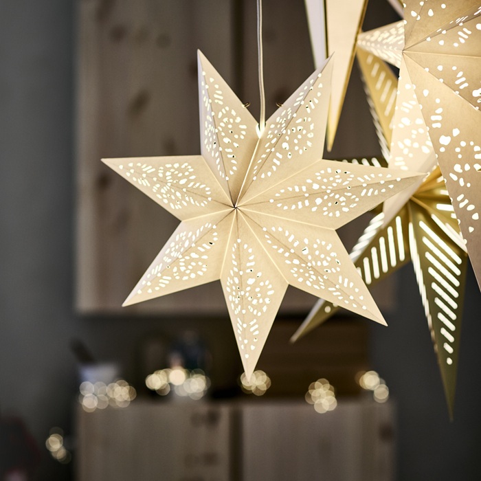 Bright STRÅLA stars hang from the ceiling in a dimly lit room with light wood-toned furniture and gray walls.