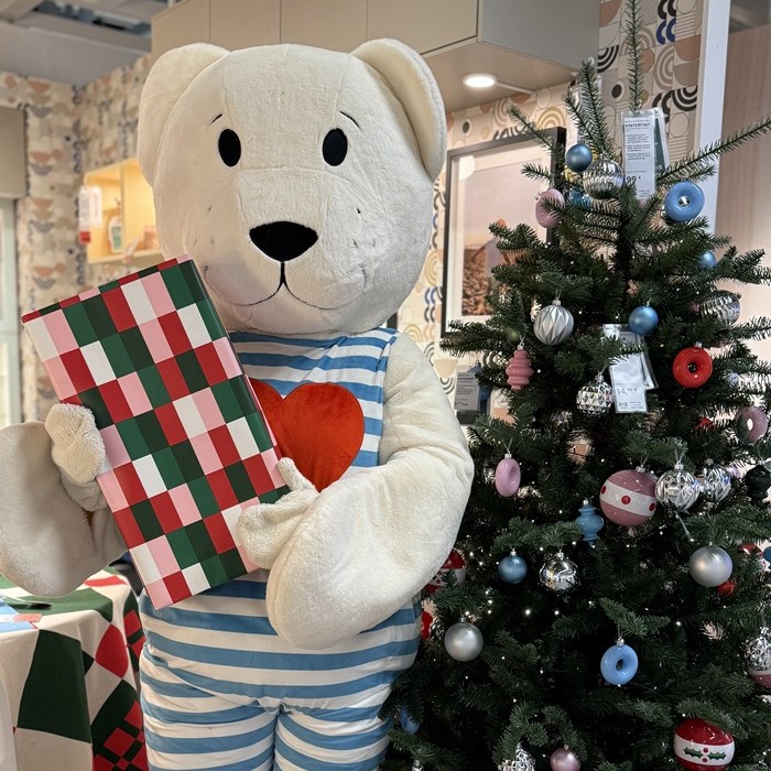 Liels balts rotaļu lācis ar iepakotu dāvanu rokās un fonā izdekorēta Ziemassvētku eglīte.  