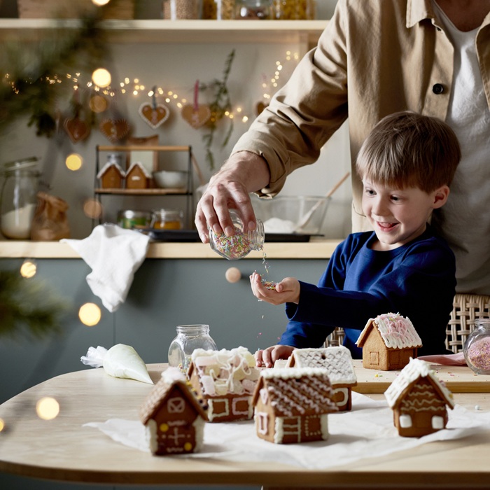Zēns zilā džemperī pie galda, uz kura atrodas vairākās ar glazūru dekorētas piparkūku mājiņas. Pieaugušais zēnam saujā ber cukura pērlītes no stikla trauka.