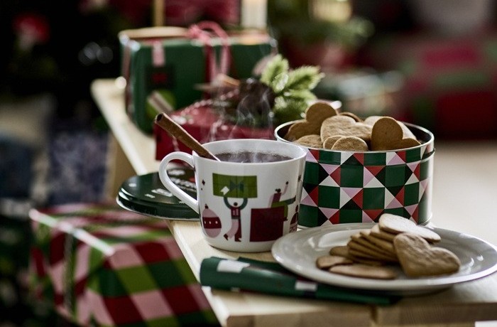 Uz sola uzklāts improvizēts galds: krūze ar kūpošu dzērienu, šķīvis ar piparkūku sirdīm, piparkūku kārba, fonā dāvanas un aizdegtas sveces.