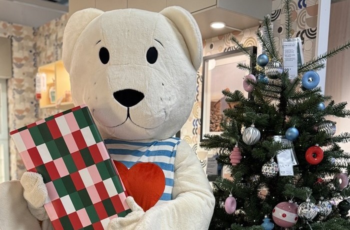 Liels balts rotaļu lācis ar iepakotu dāvanu rokās un fonā izdekorēta Ziemassvētku eglīte.  