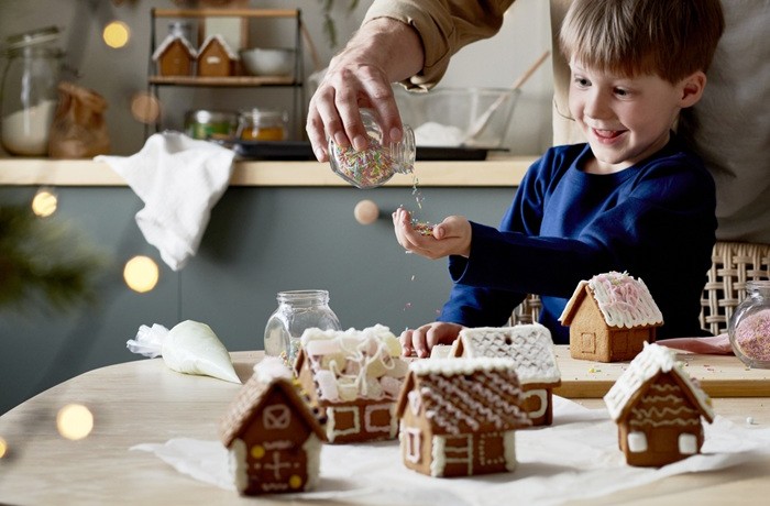 Zēns zilā džemperī pie galda, uz kura atrodas vairākās ar glazūru dekorētas piparkūku mājiņas. Pieaugušais zēnam saujā ber cukura pērlītes no stikla trauka.