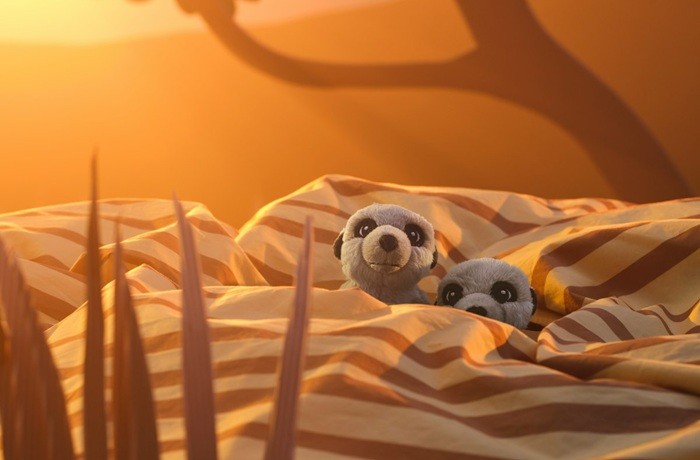 Divas mīkstās mantiņas – surikati paslēpušies svītrainā gultas veļā, tos apspīd silta gaisma.