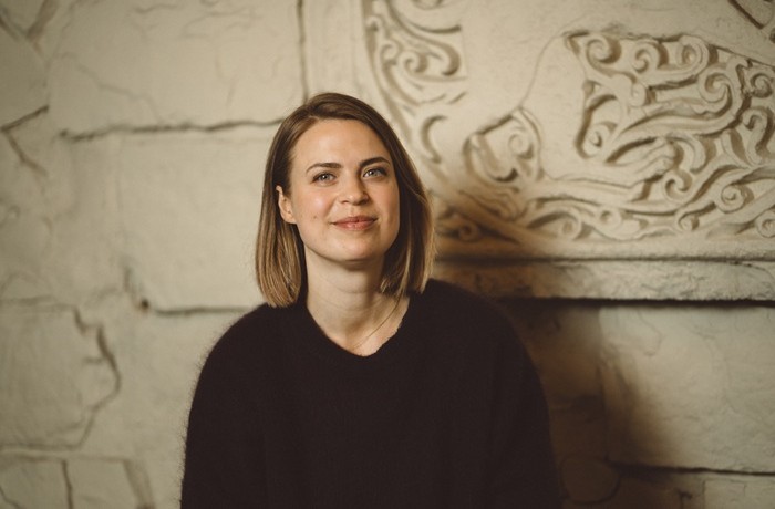 Uz gaišas akmens sienas fona redzama, melnā džemperī tērpusies, smaidīga sieviete vidēji gariem, brūniem matiem.