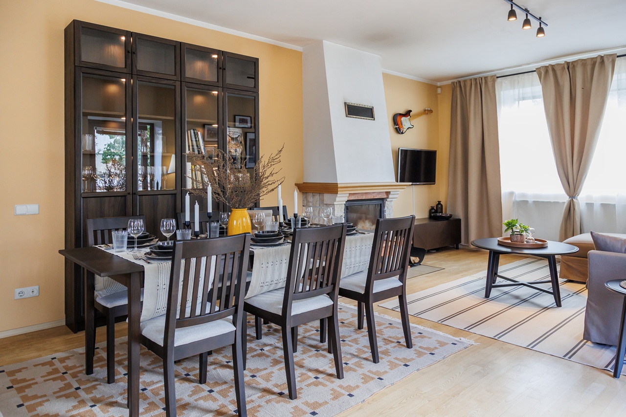 В светлой гостиной стоят обеденный стол со стульями и книжный шкаф; на заднем плане видны камин и окно со шторами.