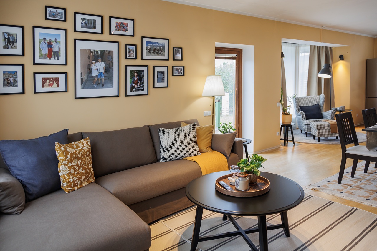 В углу гостиной — коричневый трёхместный диван с узорными подушками и круглый чёрный журнальный столик. На стене над диваном висит коллаж из картин в рамках.