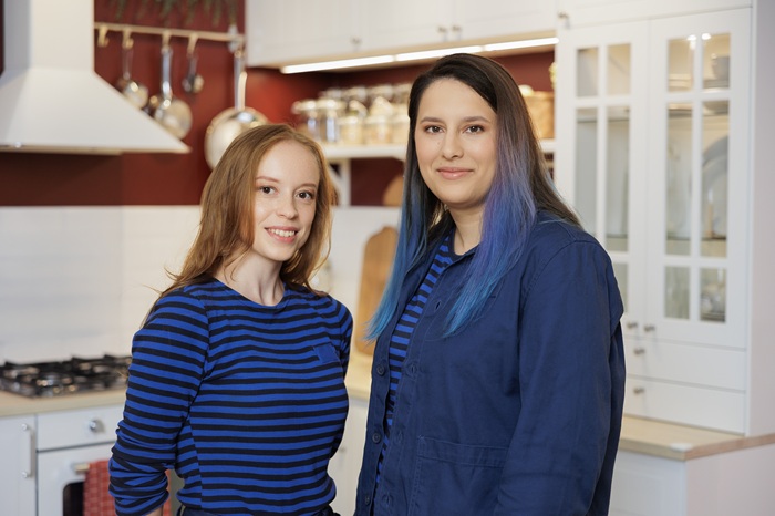 Двое дизайнеров интерьера IKEA в синей полосатой униформе уверенно стоят в выставочном зале кухни с белыми шкафами и красными стенами.