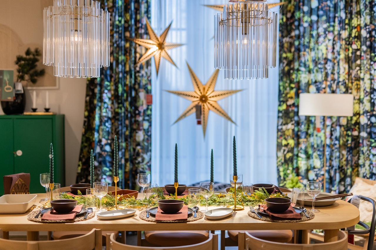 A festive dining table decorated with green candles, greenery and modern tableware, with large golden star ornaments hanging in front of patterned curtains.