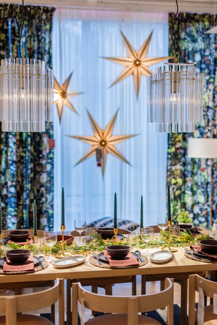 A decorated dining table with green candles and natural greenery, placed near large golden star decorations hanging in the window.