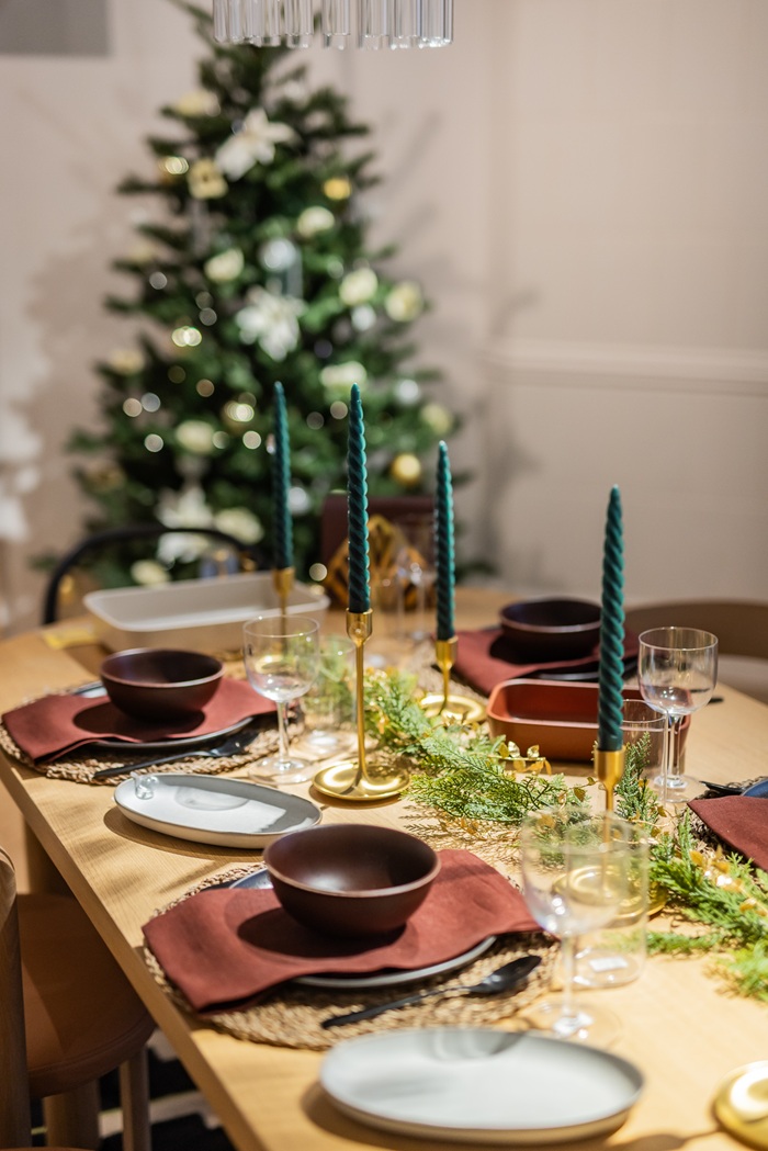 A Christmas tree stands behind a festive dining table decorated with greenery, dark-toned plates and green candles.