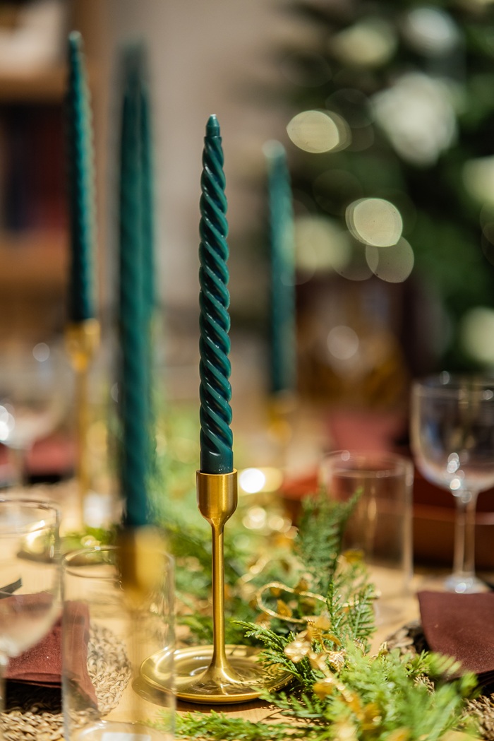 Close-up of tall twisted green candles in gold holders on a festive table, with blurred Christmas lights in the background.
