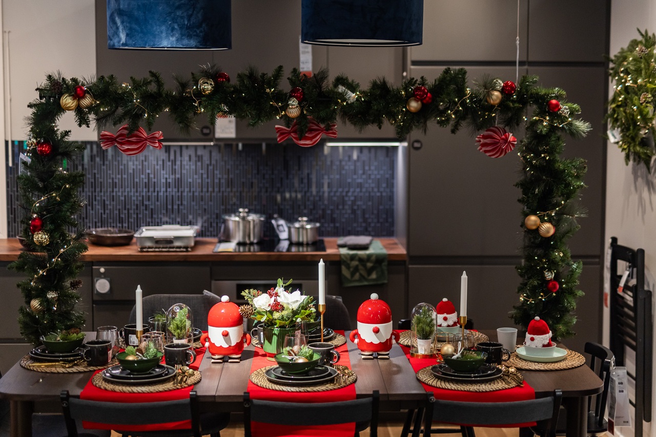 A festive dining table decorated with red and green Christmas accents, set beneath an arch of garlands with ornaments in a modern kitchen.