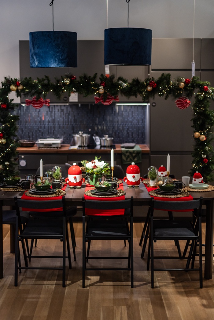 A long dining table set for Christmas with garlands overhead, red place settings and candlelight, arranged in a contemporary kitchen space.