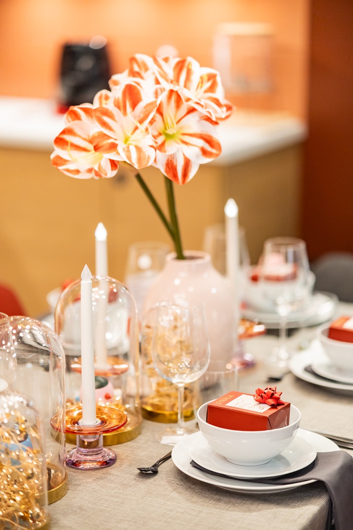 A close-up of a holiday table centerpiece featuring pink amaryllis in vases, candles and decorative glass domes filled with golden accents.