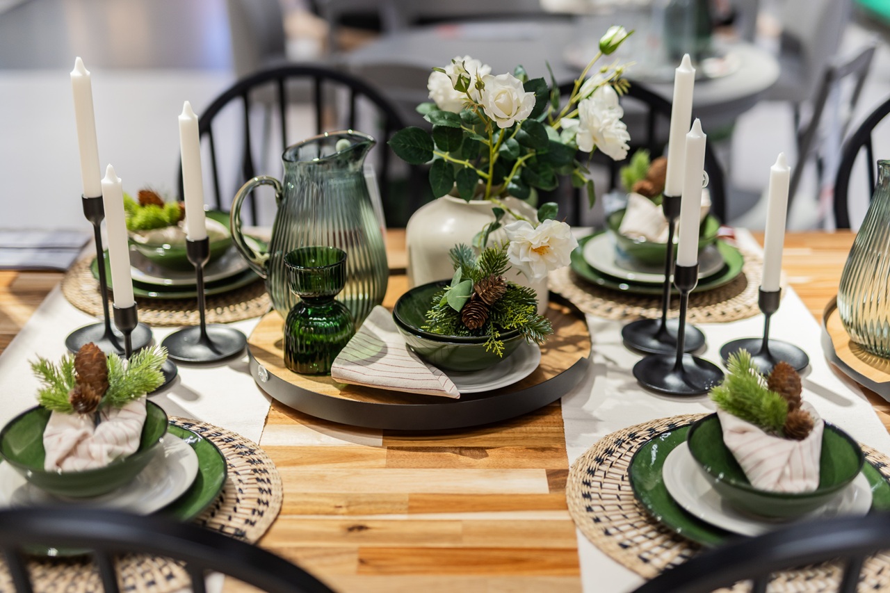 A festive dining table decorated with green plates, white flowers, tall candles and natural elements like pinecones and moss, arranged on a wooden tabletop.
