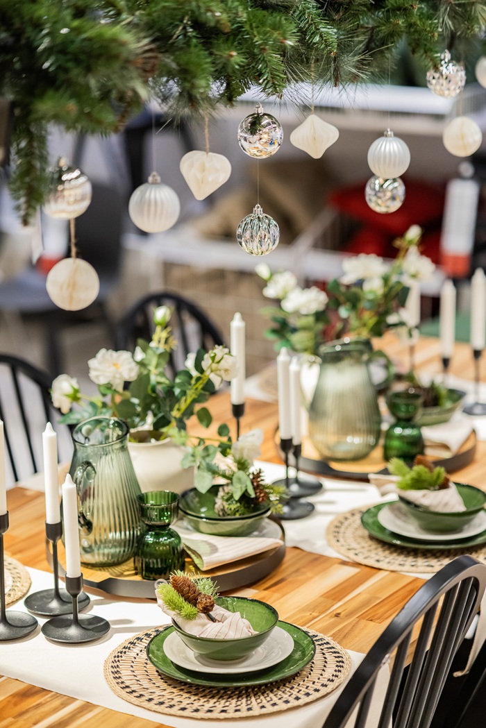 A decorated dining table with green tableware, floral arrangements and candles, displayed under hanging festive ornaments.
