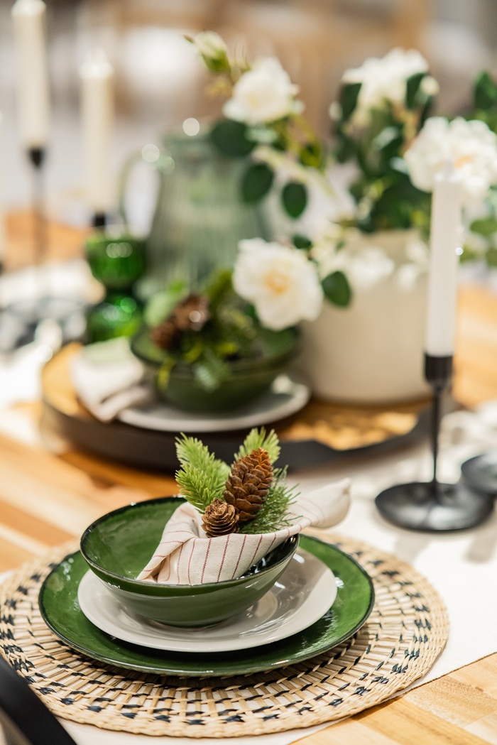 Close-up of a festive place setting with green plates, a folded napkin, pinecones and white flowers arranged as a natural decorative accent.