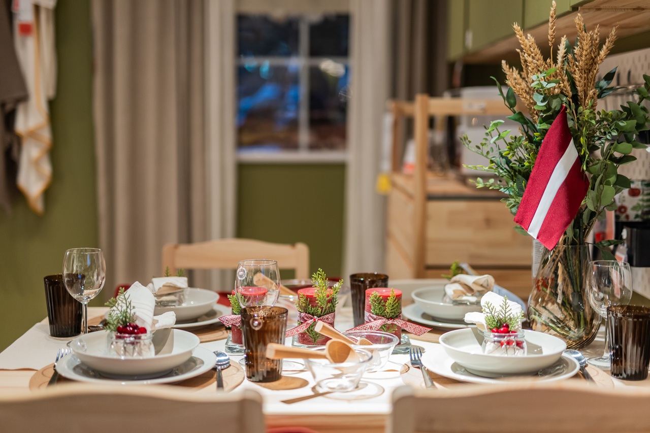 A festive dining table decorated with neutral tableware, greenery, red accents and a floral arrangement featuring the Latvian flag in a cosy dining space.