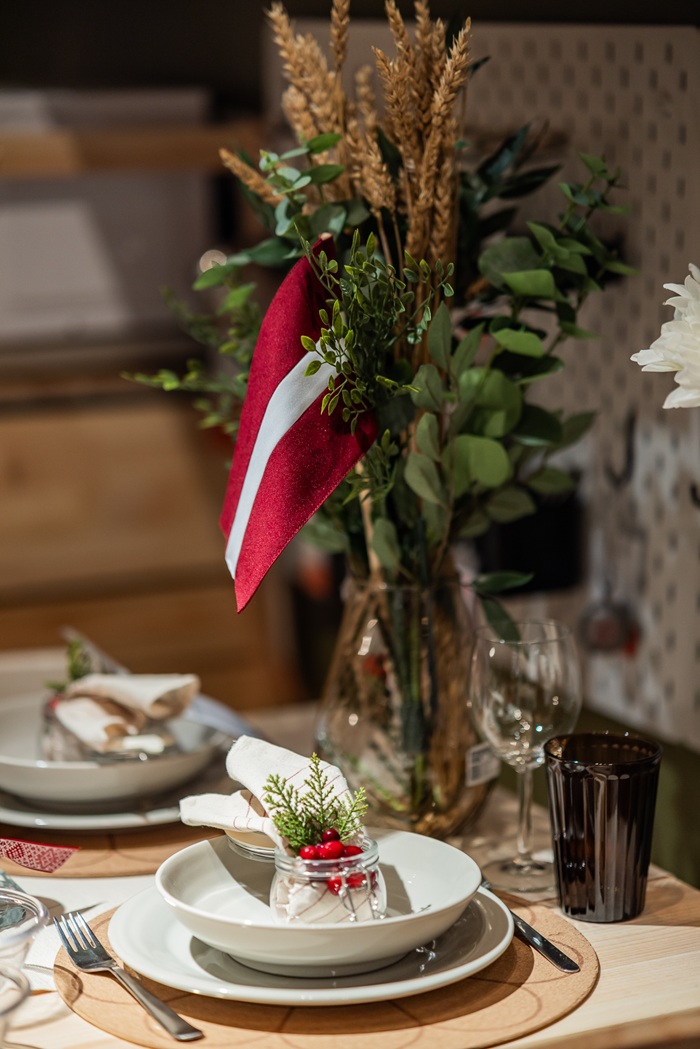 A decorated dinner table with layered plates, festive greenery, red napkin details and a Latvian flag arrangement placed at the centre.