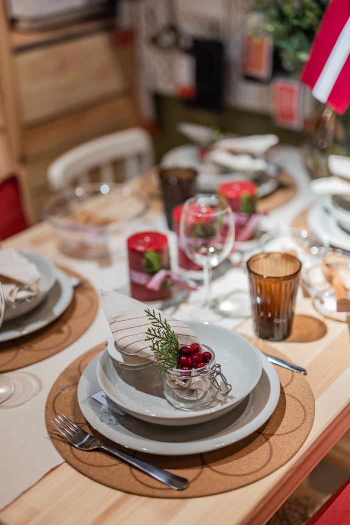A close-up of a festive place setting with white plates, greenery, red berries and a napkin tied with a ribbon in Latvian flag colours.