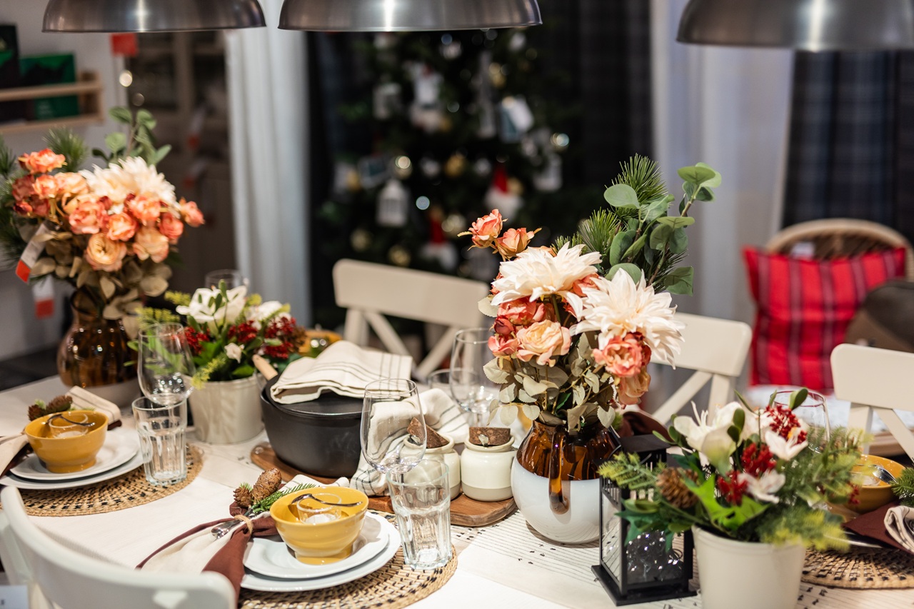 A festive dining table decorated with warm-toned floral arrangements, ceramic tableware, candles and rustic natural accents, with a Christmas tree in the background.