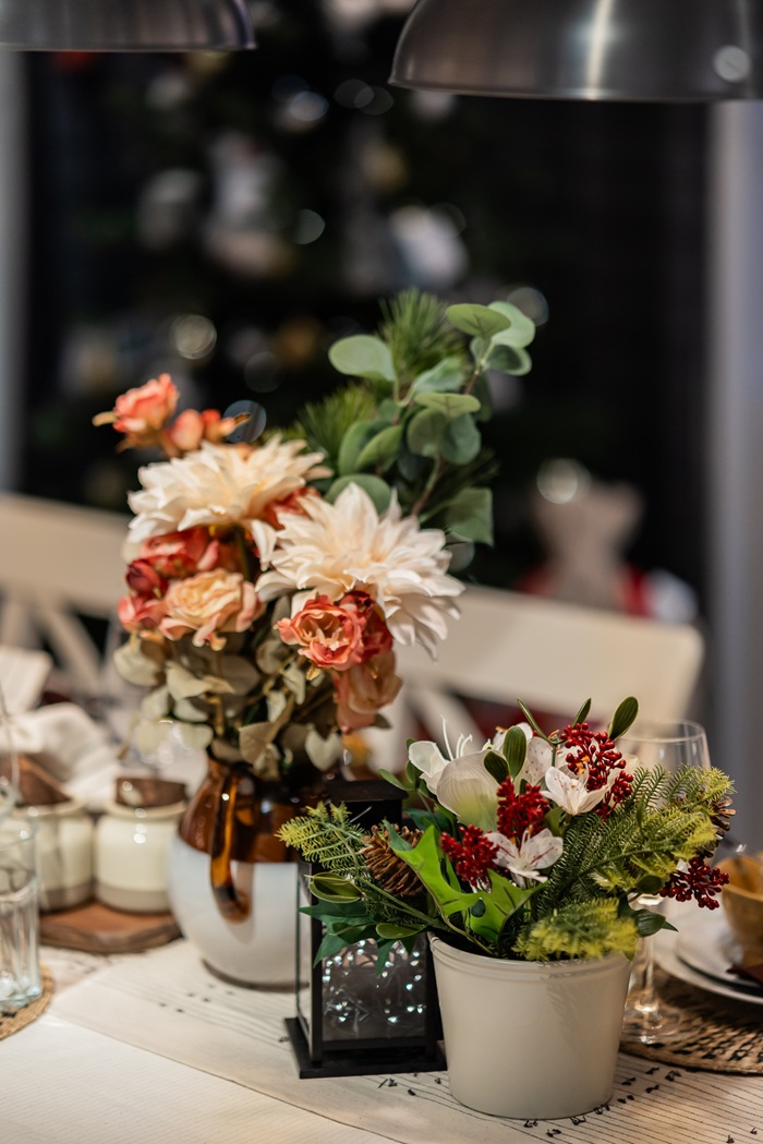 A close-up of festive table décor featuring flowers, greenery, a lantern-style candle holder and ceramic dishes arranged on a white tablecloth.