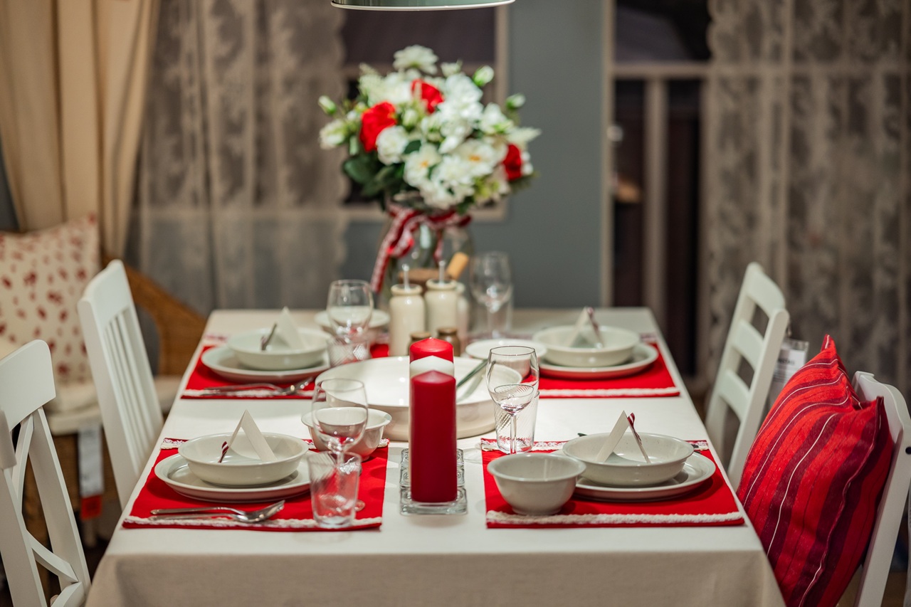 A festive dining table set with red placemats, white tableware, candles and a central bouquet of red and white flowers in a cosy dining room.