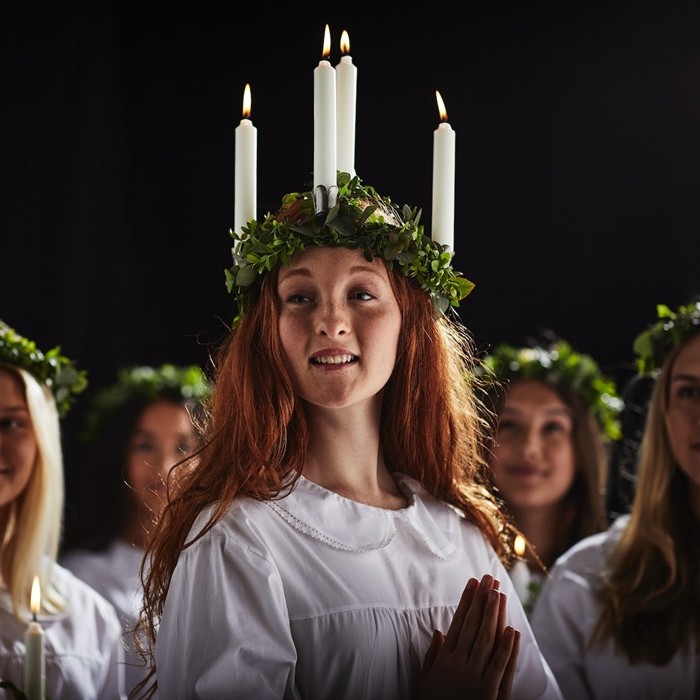 Young women dressed in white robes with wreaths of greenery on their heads take part in a St. Lucia choir celebration. One of them is wearing a crown of candles.