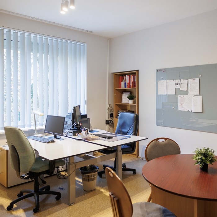 Krizių centro priimamasis iki pokyčių: prie lango stovi biuro stalai su kėdėmis, ant sienos kabo skelbimų lenta, šalia matosi apvalus medinis stalas, tačiau baldai nedera tarpusavyje.