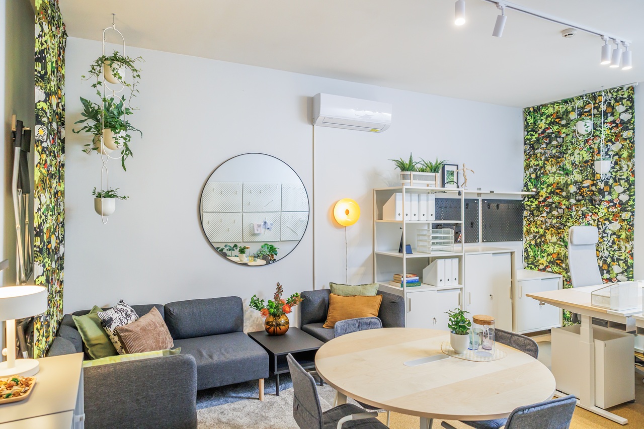 Krizių centro priimamasis po atnaujinimo: jaukiai apšviesta erdvė su sofa, baltais ir šviesaus medžio baldais, veidrodžiu bei augalais.