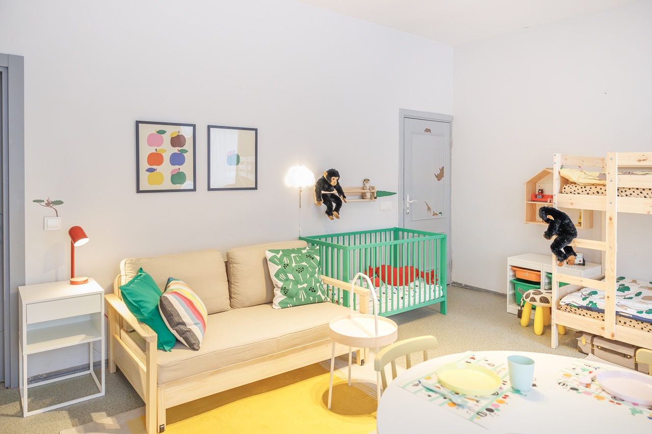 Skandinaviško stiliaus šeimos kambaryje stovi sofa-lova, apvalus stalas su kėdėmis, žalios spalvos kūdikio lovelė. Kambarys dekoruotas pagalvėlėmis, kilimais, minkštaisiais žaislais ir paveikslais.