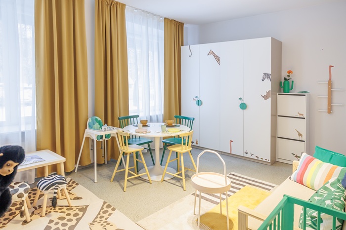 Dar viena šeimos kambario pusė: prie lango su geltonomis užuolaidomis stovi stalas įvairioms veikloms ir didelė balta spinta.
