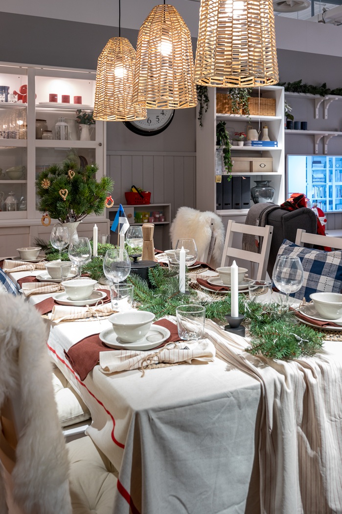 Vaade valge linaga peolauale. Sooja kumaga rippvalgustid tõstavad esile valgeid taldrikuid, rohelisi oksi ja punaseid salvrätte.        