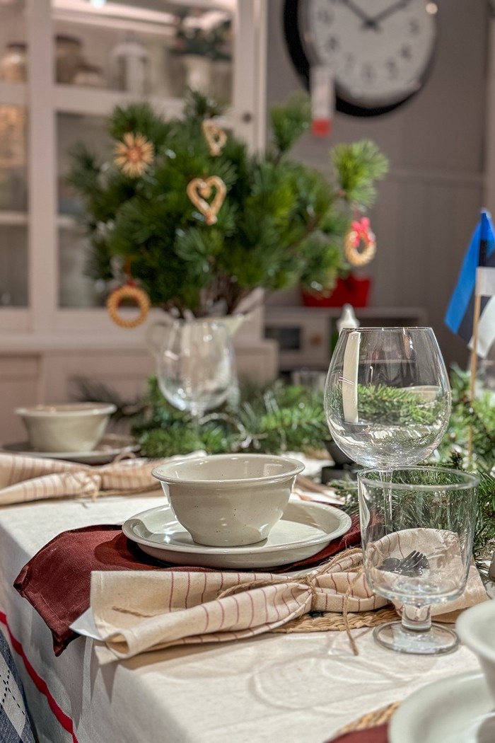 Lähivaade kaetud lauale, millel on valged taldrikud ja kirkad klaasid. Punakaspruun salvrätik annab värvikontrasti ja taustal ripuvad rohelistel okstel õlgedest kaunistused.                                