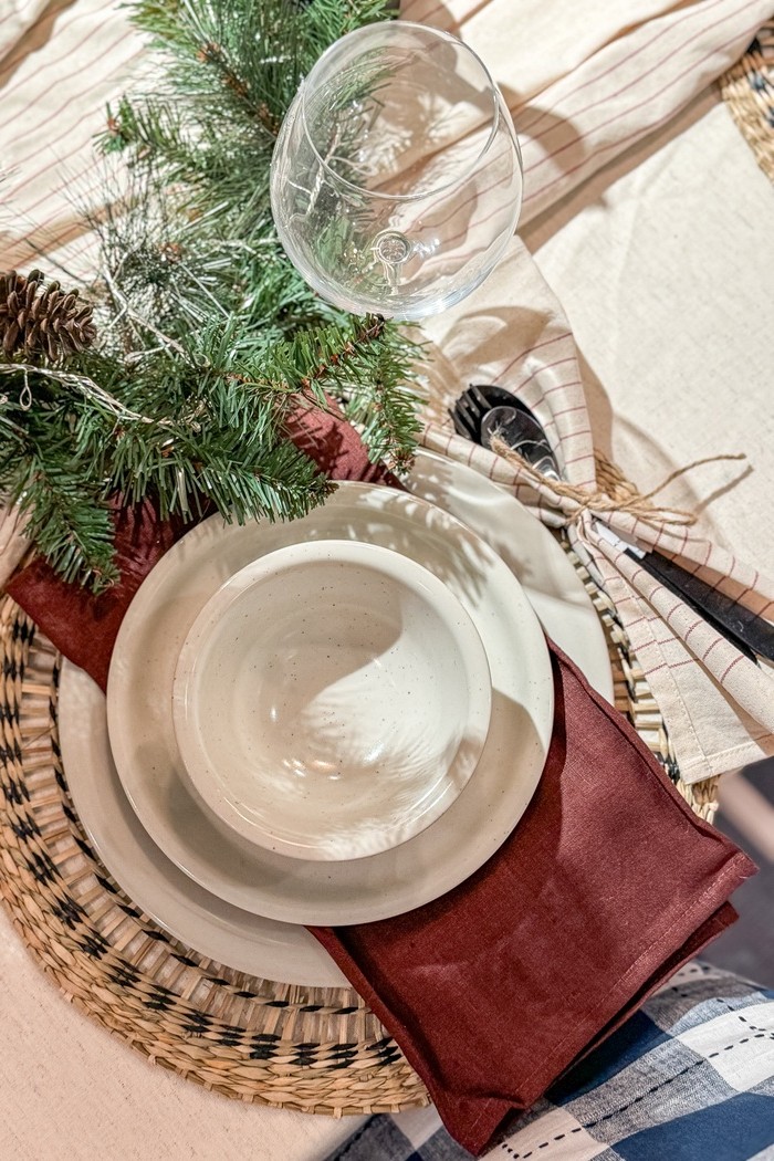 Ülaltvaade valgele kausile ja taldrikutele, mis on laotud virna. Alumisel taldrikul on pruunikaspunane salvrätik ja taldriku kõrval on roheline oks käbiga.